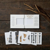 On a dark wooden surface, an open white envelope displays decorative cards with intricate black and brown designs, complemented by tall dried grass. The scene evokes subtle Viking motifs and a nod to Danish history. Product: Card Pack - Viking Finds Square Card Folder by Koustrup & Co, Denmark.