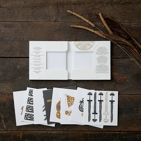 On a dark wooden surface, an open white envelope displays decorative cards with intricate black and brown designs, complemented by tall dried grass. The scene evokes subtle Viking motifs and a nod to Danish history. Product: Card Pack - Viking Finds Square Card Folder by Koustrup & Co, Denmark.