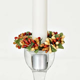 A white taper candle in a clear glass holder surrounded by an autumn-colored pip berry and green leaf ring.