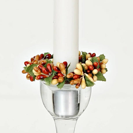 A white taper candle in a clear glass holder surrounded by an autumn-colored pip berry and green leaf ring.