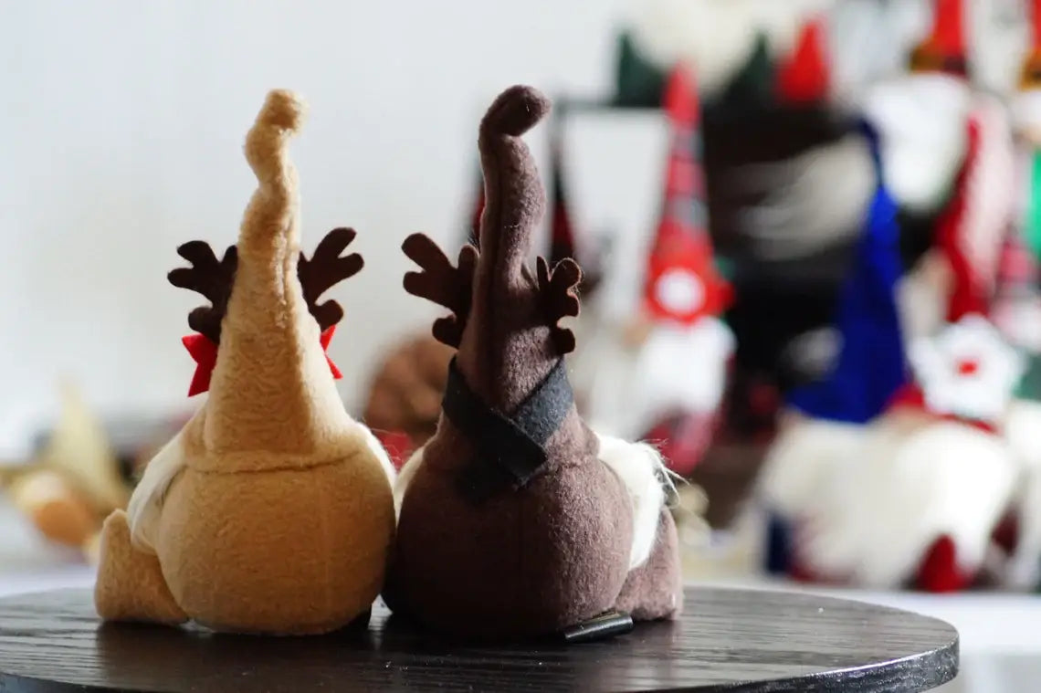 Two Rudolph Gnomes with pointy hats and antlers, embodying whimsical Christmas characters, sit on a round wooden surface, facing away. A blurred background reveals more Rudolph Gnomes in various colors.