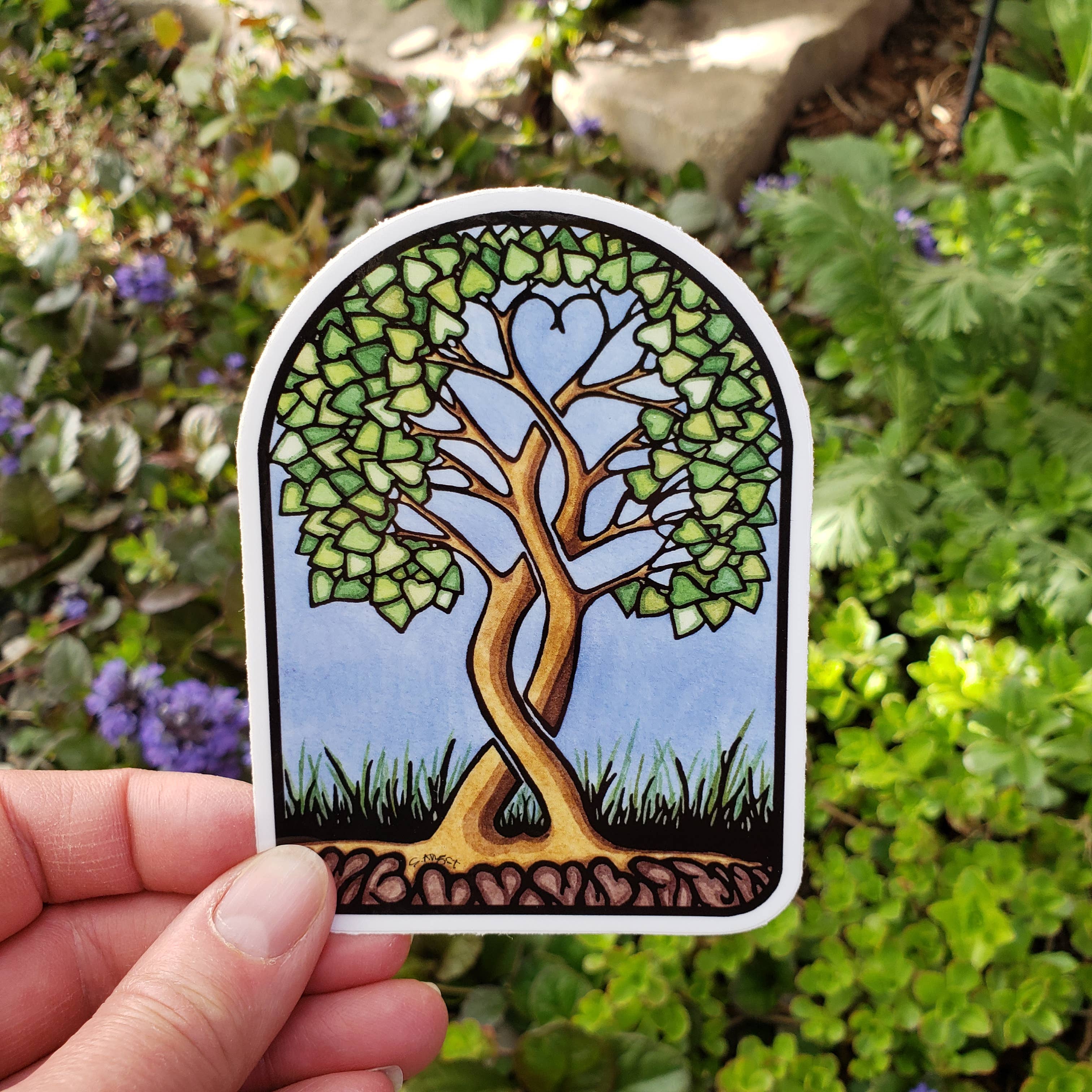 A hand showcases the Together Forever Vinyl Sticker by Sarah Angst Art, featuring two heart-leaf intertwined trees against a sky backdrop with green plants and flowers. This enchanting piece is UV protected and weatherproof for durability.