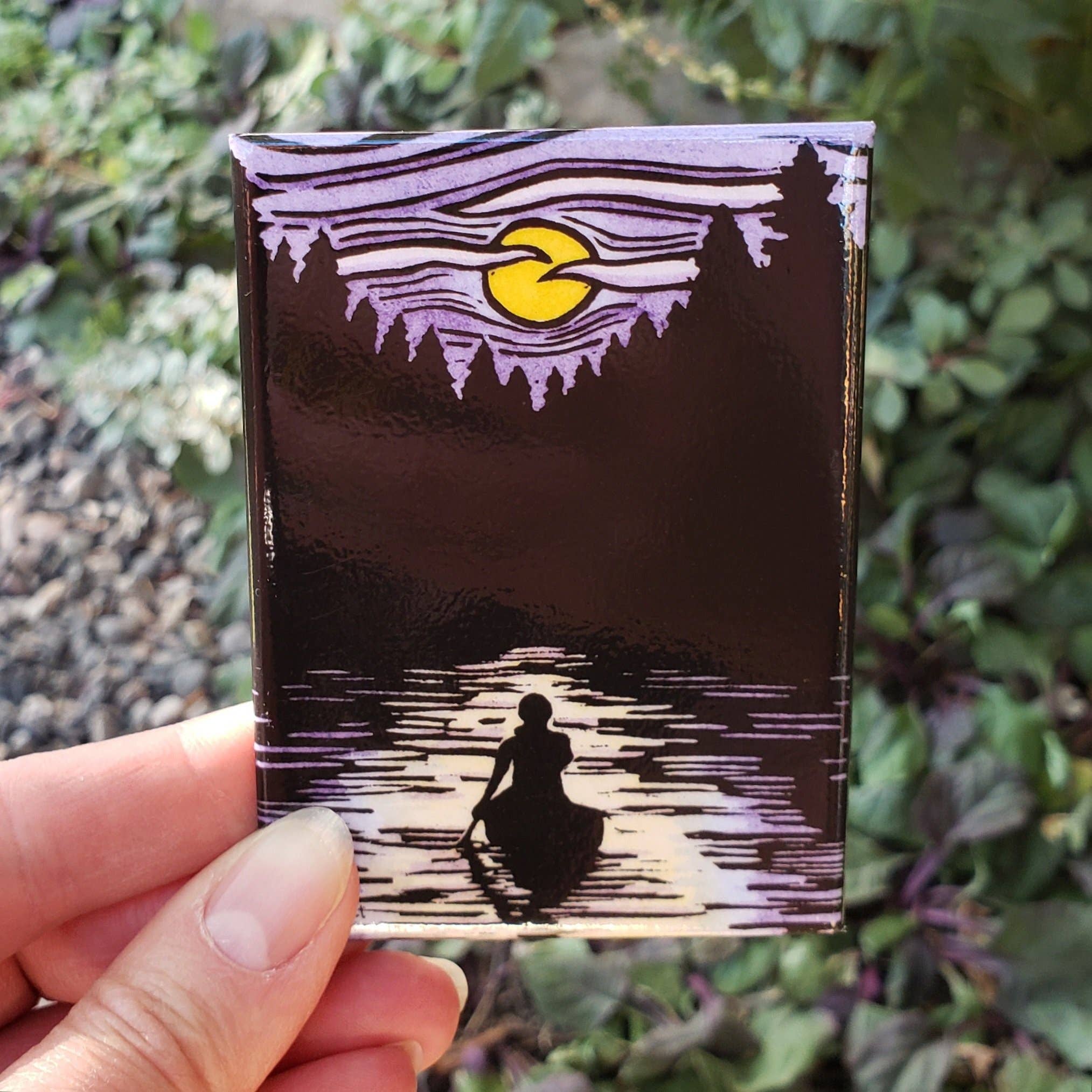 A hand holds the Magnet - Solitude Canoe Magnet, depicting a nighttime canoe gliding on a serene lake under a purple sky and the luminous yellow moon, surrounded by tree silhouettes.