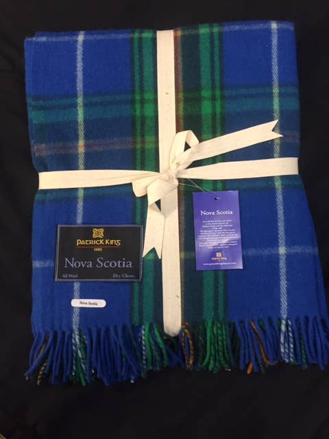 A folded blue, green, and white Nova Scotia Deluxe Highland Tartan Blanket - Merino Wool is tied with a cream ribbon and features two attached product tags.