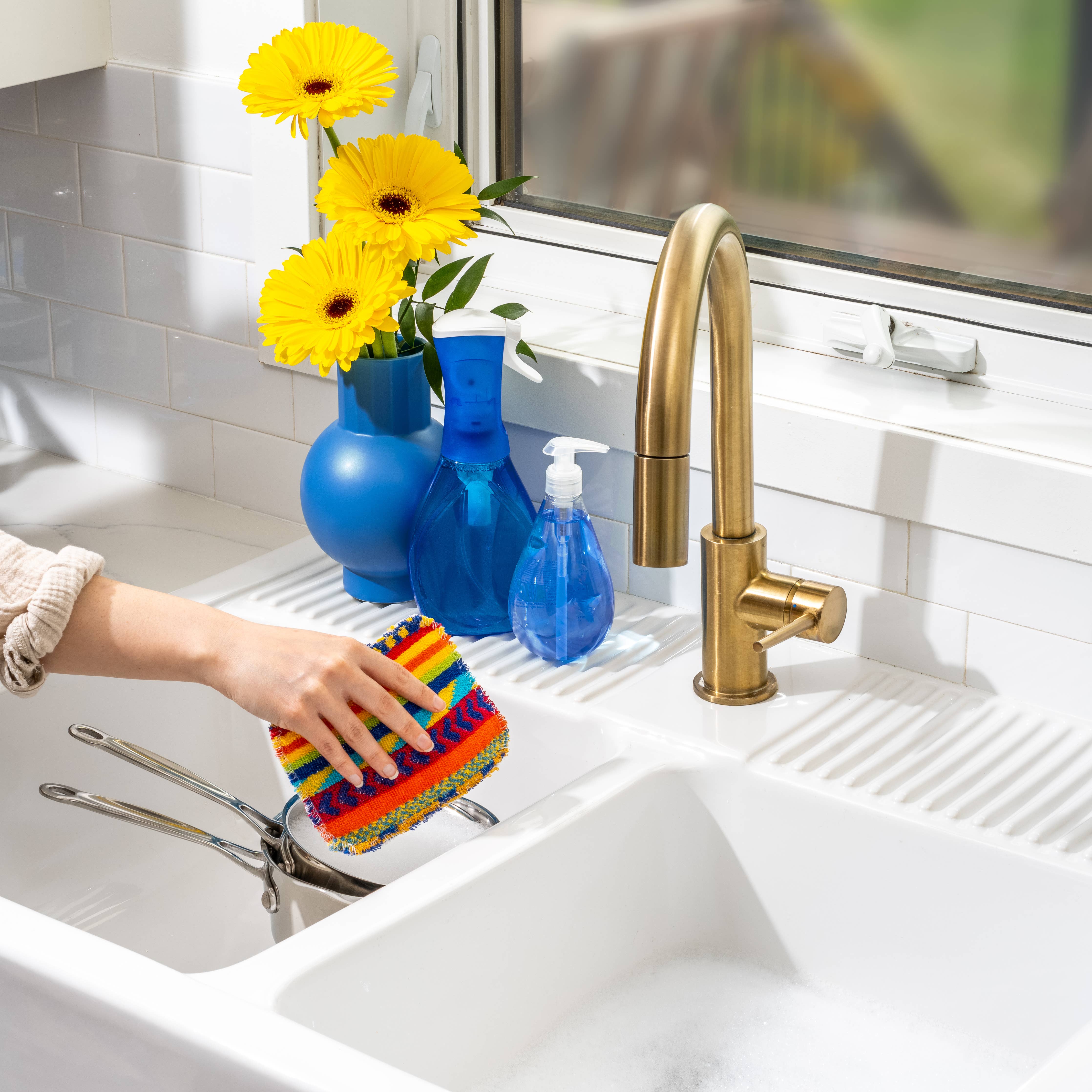 A hand uses an Euroscrubby Multi-Purpose Scrubber to wash utensils in the sink, where a brass faucet gleams. A blue vase holding yellow flowers sits on the windowsill, adding color to the kitchen.