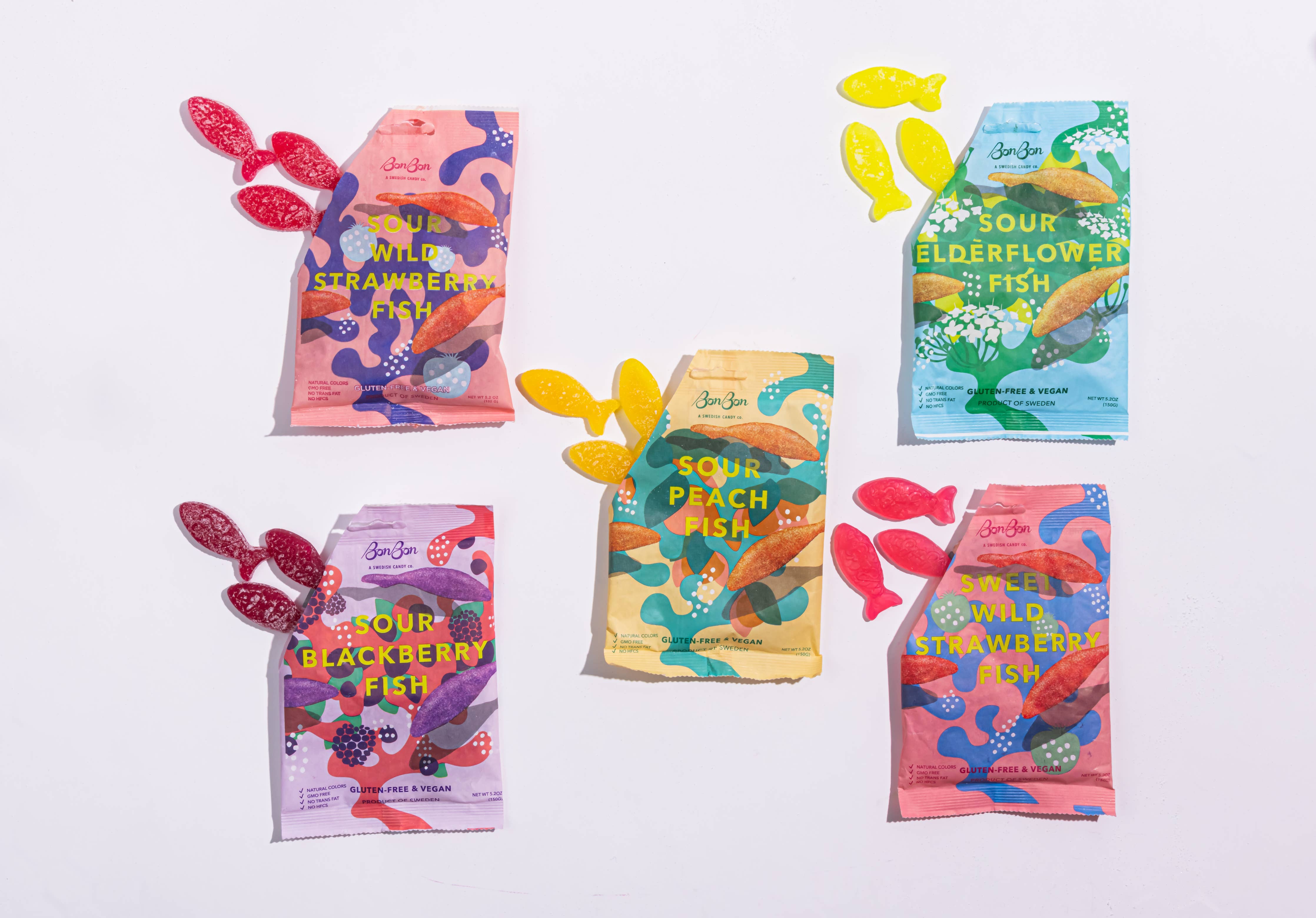 Five vibrant bags of Bon Bon candy, showcasing an array of flavors, include sour wild strawberry fish-shaped treats scattered around them, all proudly vegan and gluten-free.