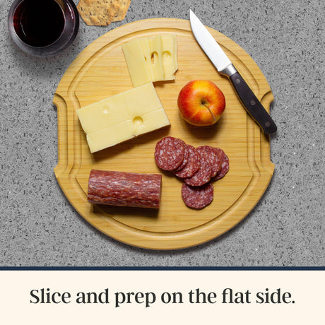 Bamboo charcuterie board featuring sliced cheese, salami, an apple, a knife, a cracker, and a glass of red wine.