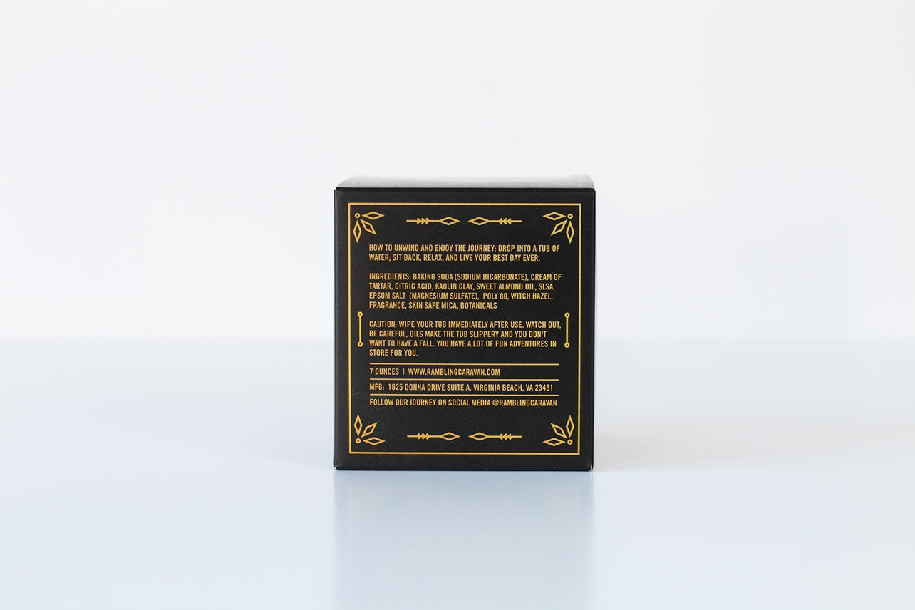 A black box with gold accents and text showing instructions, nourishing ingredients, caution, website, address, and social media for the Bath Bomb: Sandalwood & Vanilla, photographed against a plain white background.