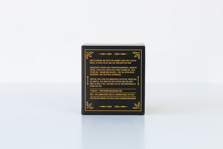 A black box with gold accents displays a label featuring product details for the Sandalwood & Vanilla Bath Bomb, including ingredients, cautions, website, and social media info on a white background.