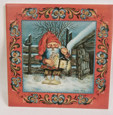 The 6x6 "Tomte with Logs and a Lantern in the Snow" tile features a gnome in red with a lantern and logs, standing in snow by a fence and cabin, framed by Norwegian rosemaling. Includes cork backing and hanger.