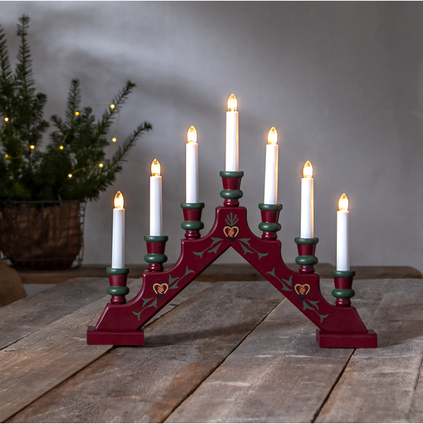 The Candlestick Sara, a wooden candelabra evoking Swedish Christmas tradition, stands proudly on a rustic table with its seven candles aglow. In the background, a small potted evergreen tree adorned with twinkling lights completes the cozy holiday scene.