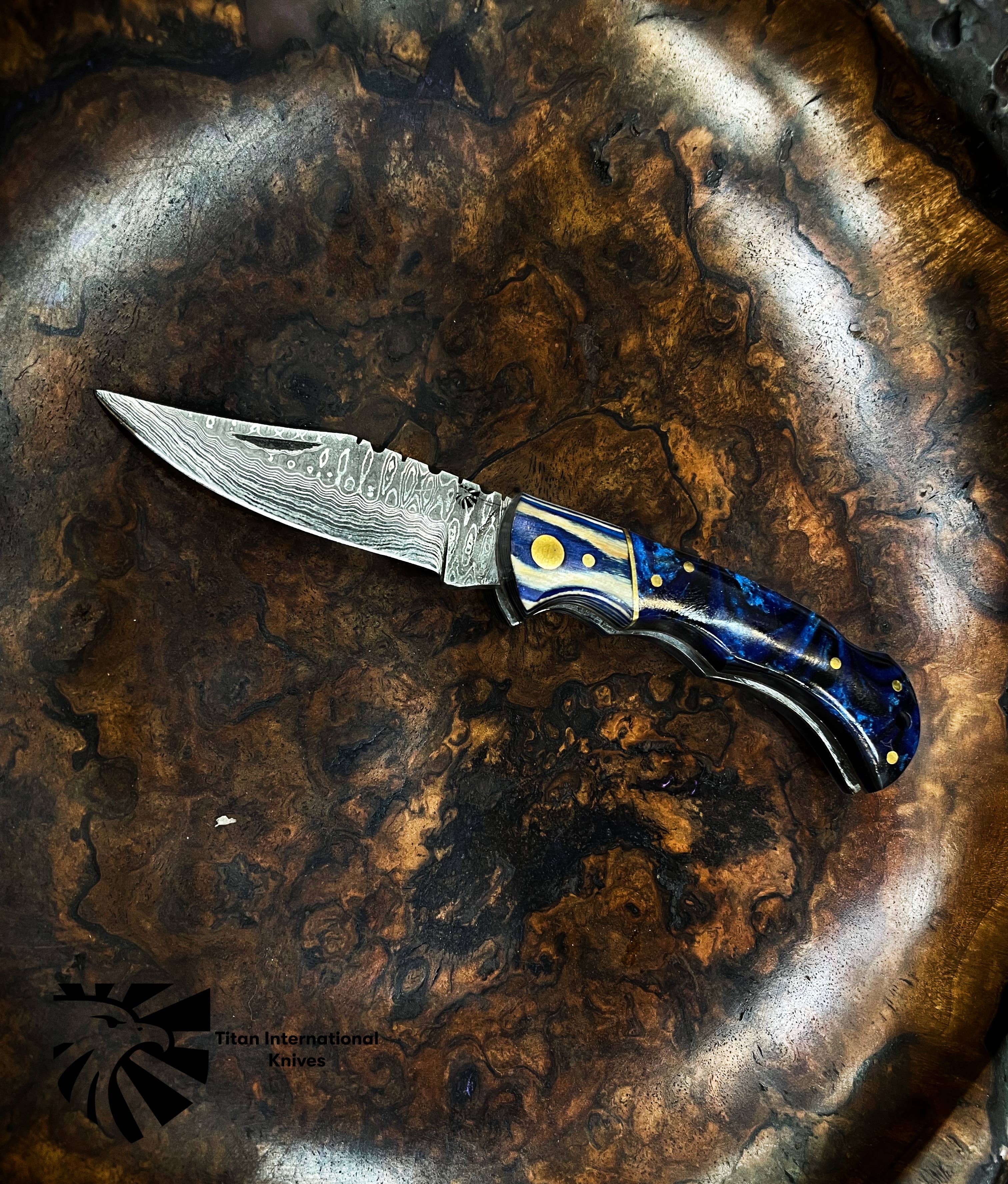 The Titan Celestial Damascus Pocket Knife features a hand-forged patterned blade and a blue handle with gold accents, displayed on a textured wooden surface.