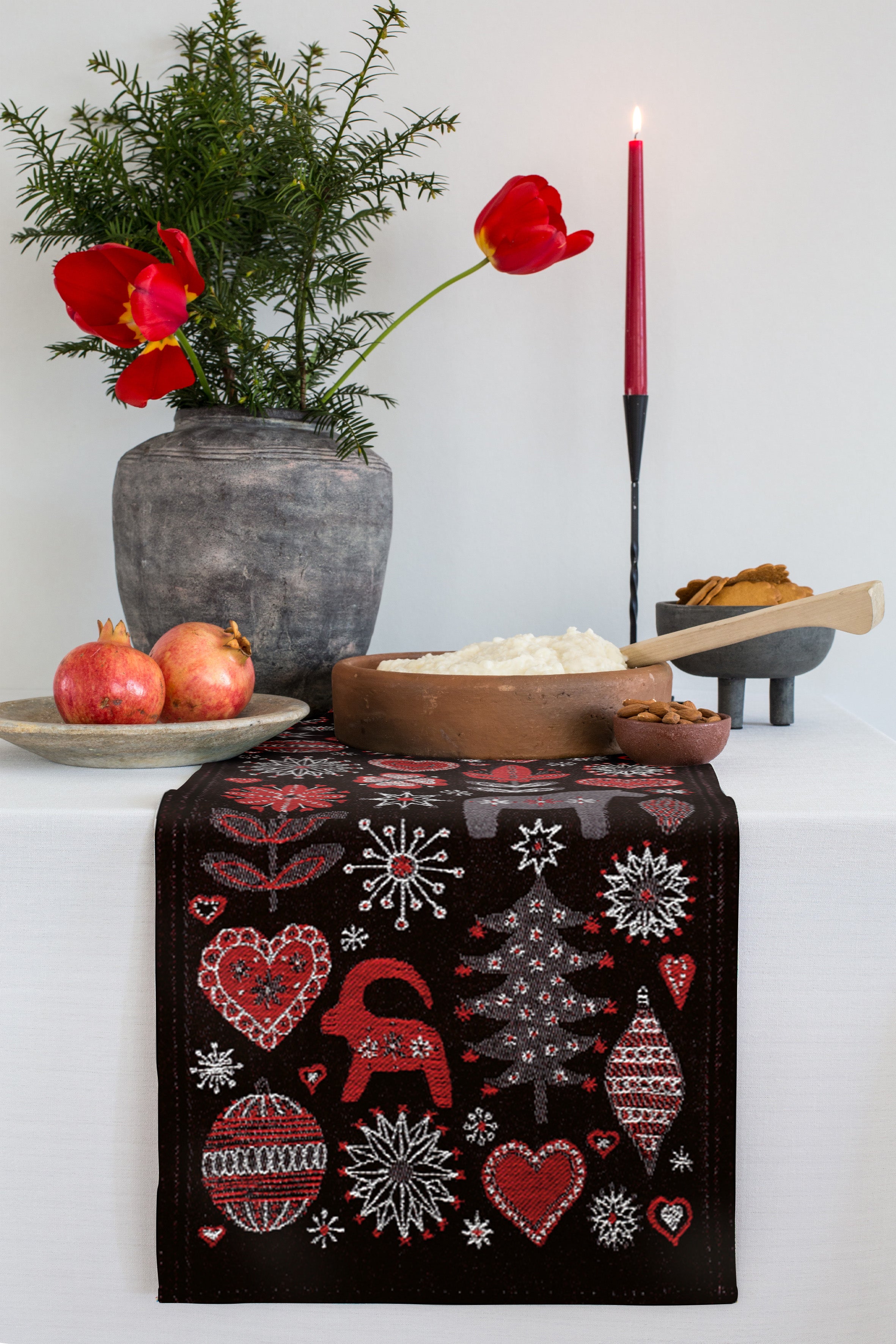 The festive table is elegantly adorned with a candle, vibrant red flowers in a vase, ripe pomegranates, and rice in a wooden bowl. Completing the scene is the Runner: Christmas Julnatt Ekelund Master Weavers, made from organic cotton in Sweden and draped gracefully with holiday-themed patterns.