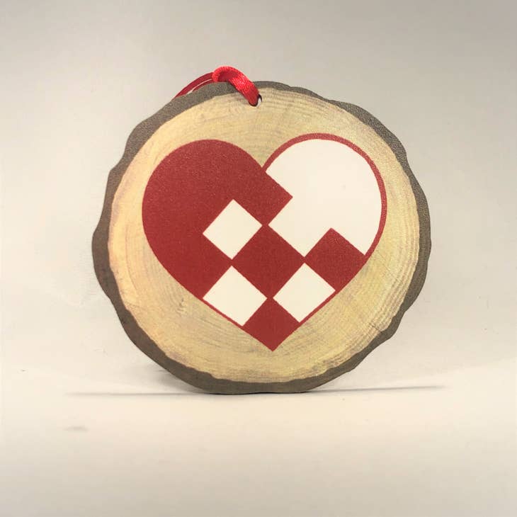 The Ornament: Pleated Heart Nordic Collection, inspired by Hans Christian Andersen's timeless tales, features a round wooden ornament adorned with a red and white heart design of interlocking geometric shapes, all elegantly suspended by a red ribbon.
