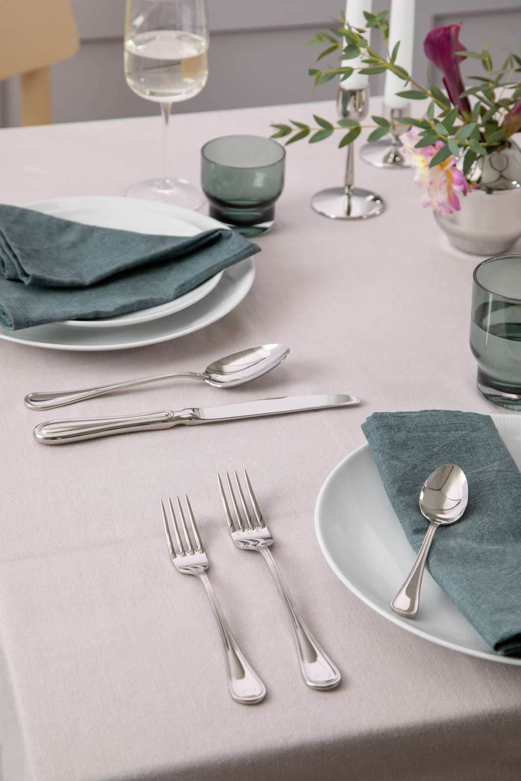 Elegant table setting with plates, teal napkins, Gense: Oxford Cutlery Set - Polished Steel, green glassware, a white wine glass, a small flower arrangement on a beige tablecloth.