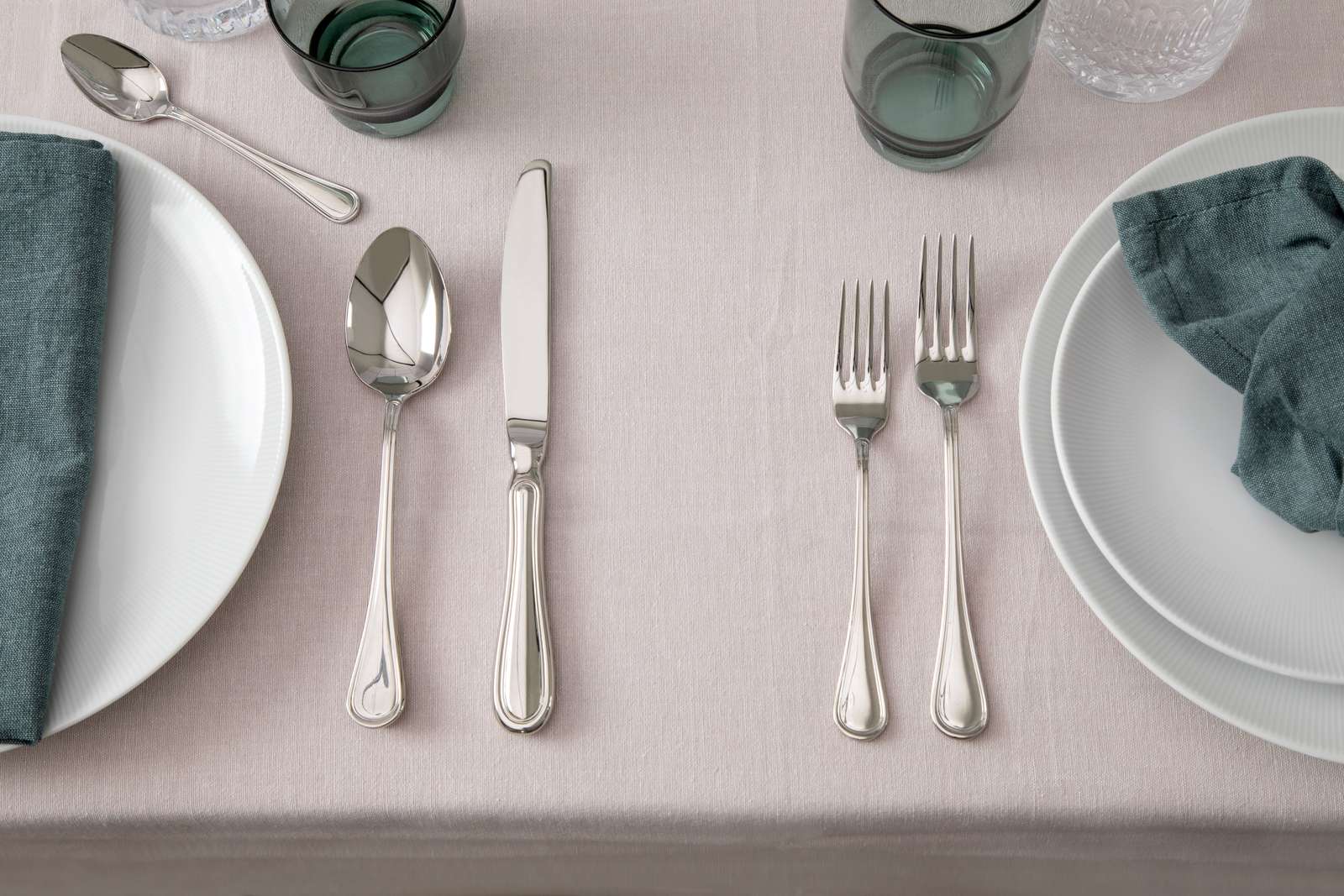 The table setting includes white plates and teal napkins on a light tablecloth, complemented by the Gense: Oxford Cutlery Set - Polished Steel, known for being dishwasher-safe. Above each plate, there are four glasses.