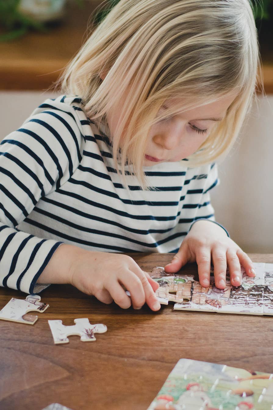 A young child with blonde hair and a striped shirt focuses on assembling the Elsa Beskow "Children of the Forest" 4 Tray Puzzles in a Wooden Box at a wooden table.