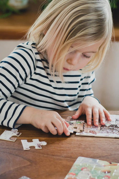 A young child with blonde hair and a striped shirt focuses on assembling the Elsa Beskow "Children of the Forest" 4 Tray Puzzles in a Wooden Box at a wooden table.