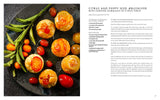 Six æbleskiver Danish pancakes topped with candied kumquats and cherries, served on a black plate with herbs and cherry tomatoes, beside the book Aebleskiver by Pim Pauline Overgaard: A New Take on Traditional Danish Pancakes.
