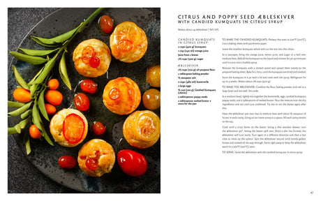 Six æbleskiver Danish pancakes topped with candied kumquats and cherries, served on a black plate with herbs and cherry tomatoes, beside the book Aebleskiver by Pim Pauline Overgaard: A New Take on Traditional Danish Pancakes.