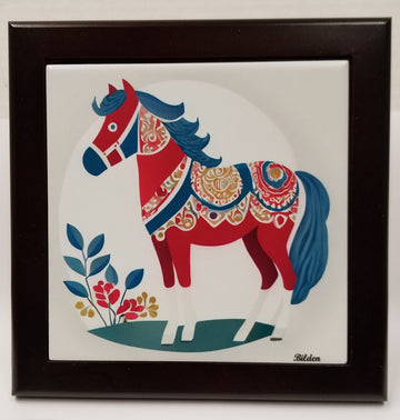 The Swedish Dala Horse Winter Folk Horse is showcased on a white tile, adorned with elaborate blue and gold designs and flanked by small plants, all enclosed in a sleek wood frame.