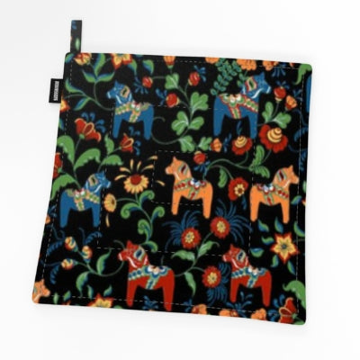 The Leksand Mini Dala Horse Potholder (9" × 9") features a black background with blue, red, and orange floral and horse motifs inspired by Scandinavian folk art—perfect as a Swedish kitchen textile.