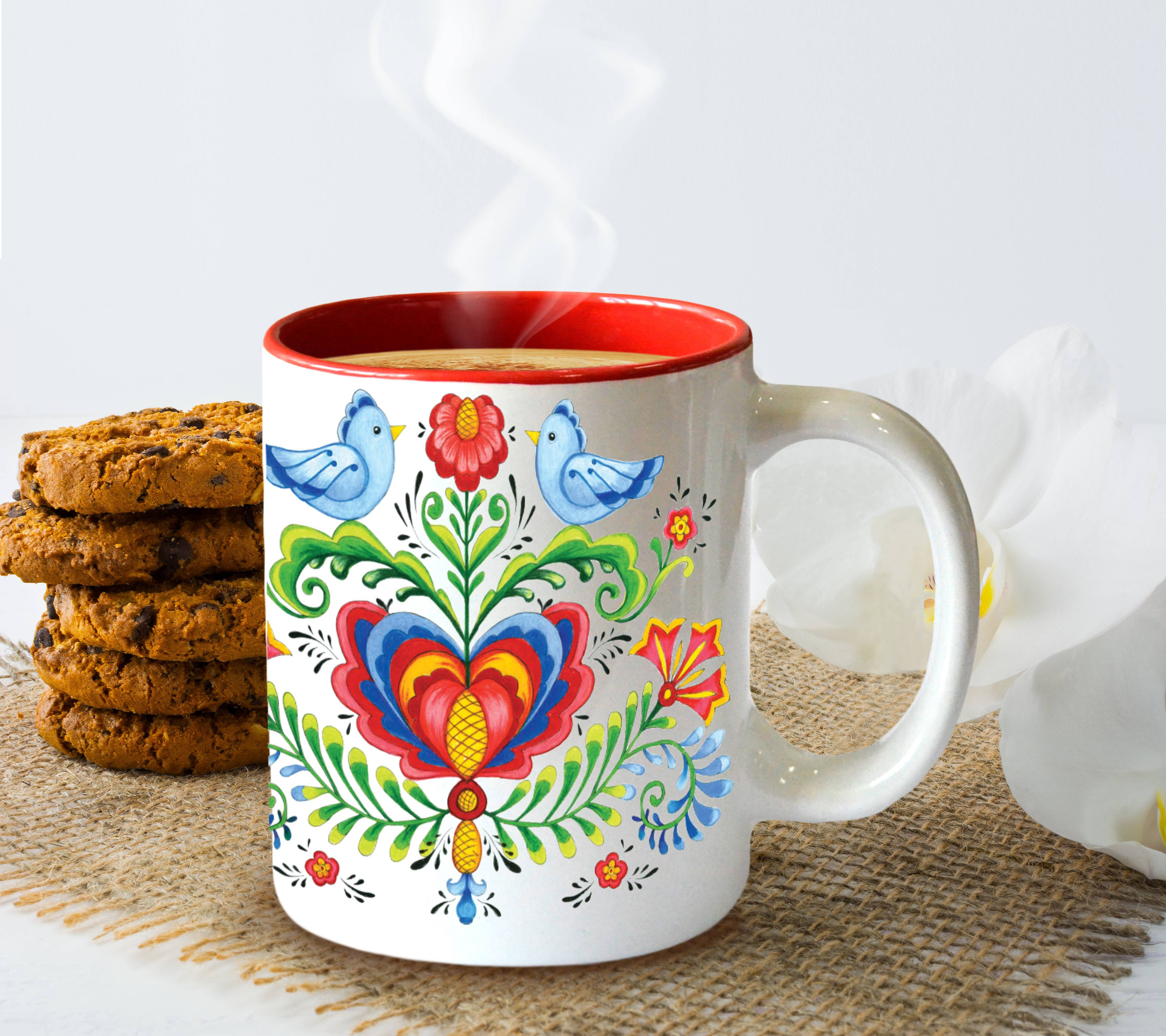 The Rosemaling & Lovebirds Ceramic Coffee Mug with Red Interior (12 oz), decorated with colorful floral and bird designs, sits filled with a hot drink on a burlap mat beside cookies and delicate white flowers.