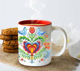 The Lovebirds Swedish Folk-Art Ceramic Coffee Mug, a white 12 oz cup with colorful bird and floral designs and a red interior, sits on burlap beside cookies and white flowers, filled with steaming coffee.