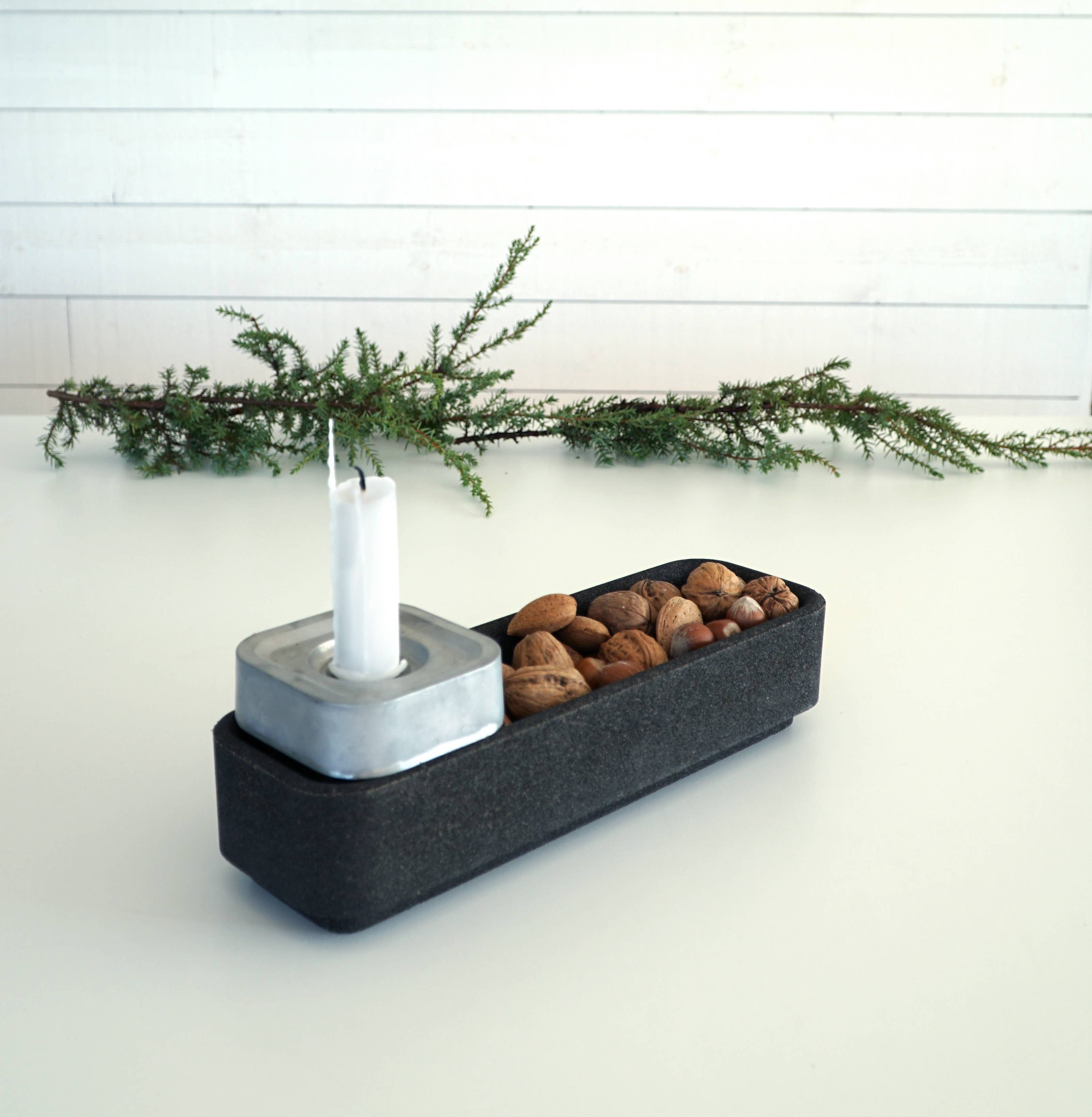 A black rectangular tray includes a Stumpastaken metal candle holder, assorted nuts, and an evergreen branch. Beside it, a recycled Corkbowl Black contributes an eco-friendly element to the scene.