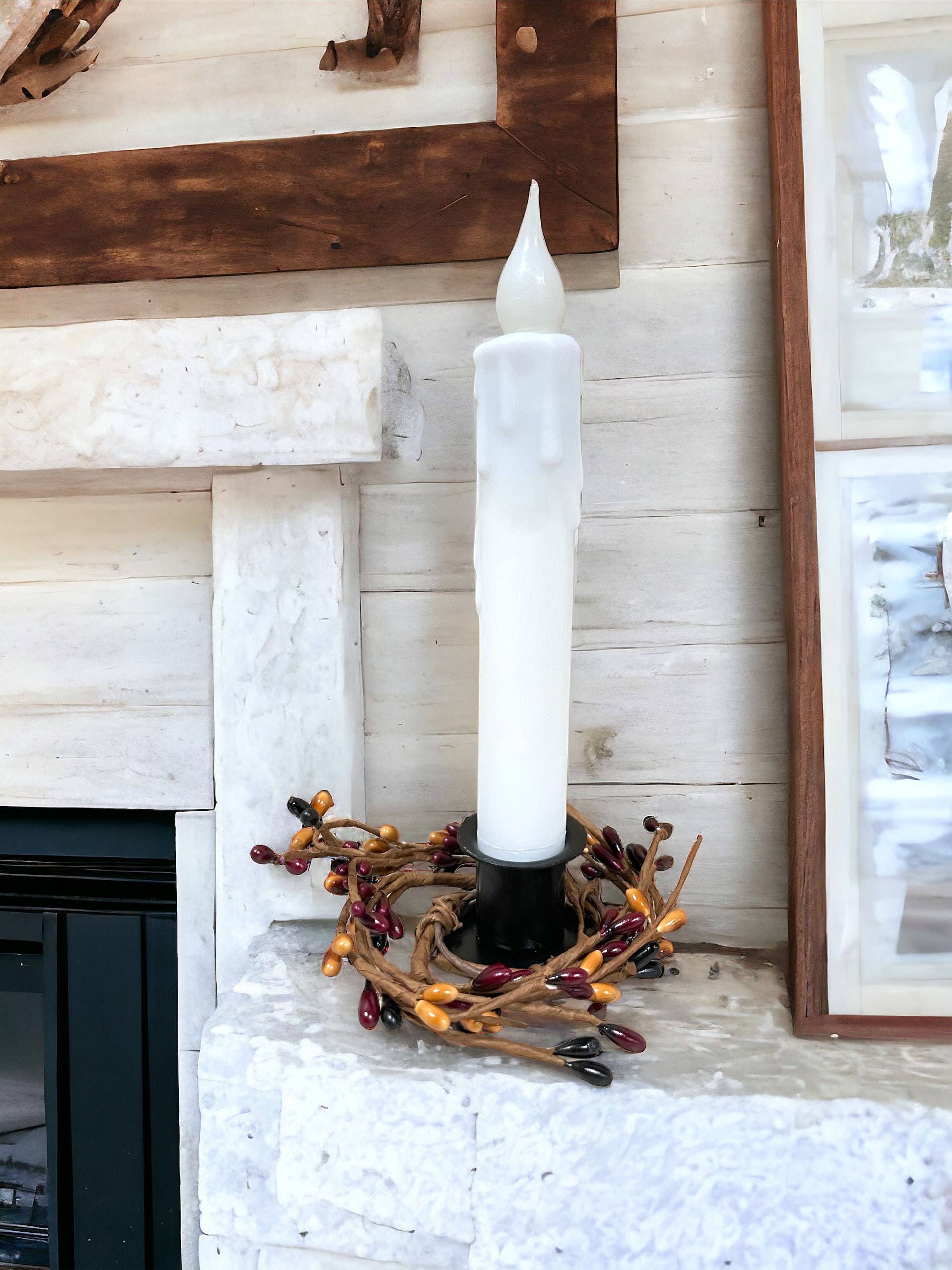 A 7-inch solid white LED battery-operated taper candle with a realistic flickering bulb in a black holder sits on a stone mantel, surrounded by a decorative wreath adorned with red, yellow, and brown beads.