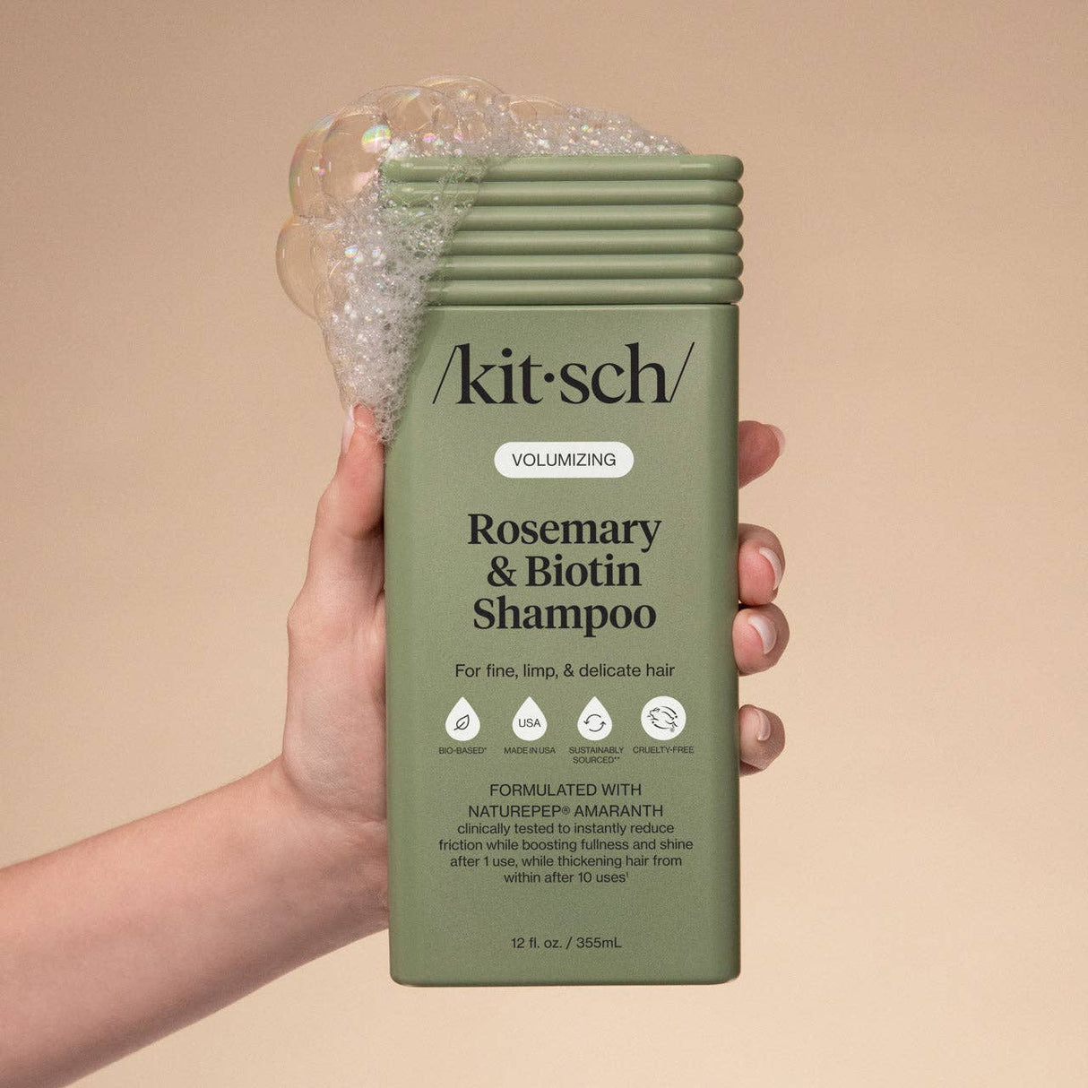 A hand holds a green bottle of Kitsch Hair: Volumizing Rosemary & Biotin Shampoo 12oz with bubbles on top; the label notes it's ideal for boosting fullness in fine, limp, and delicate hair.