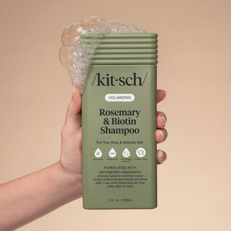 A hand holds a green bottle of Kitsch Hair: Volumizing Rosemary & Biotin Shampoo 12oz with bubbles on top; the label notes it's ideal for boosting fullness in fine, limp, and delicate hair.