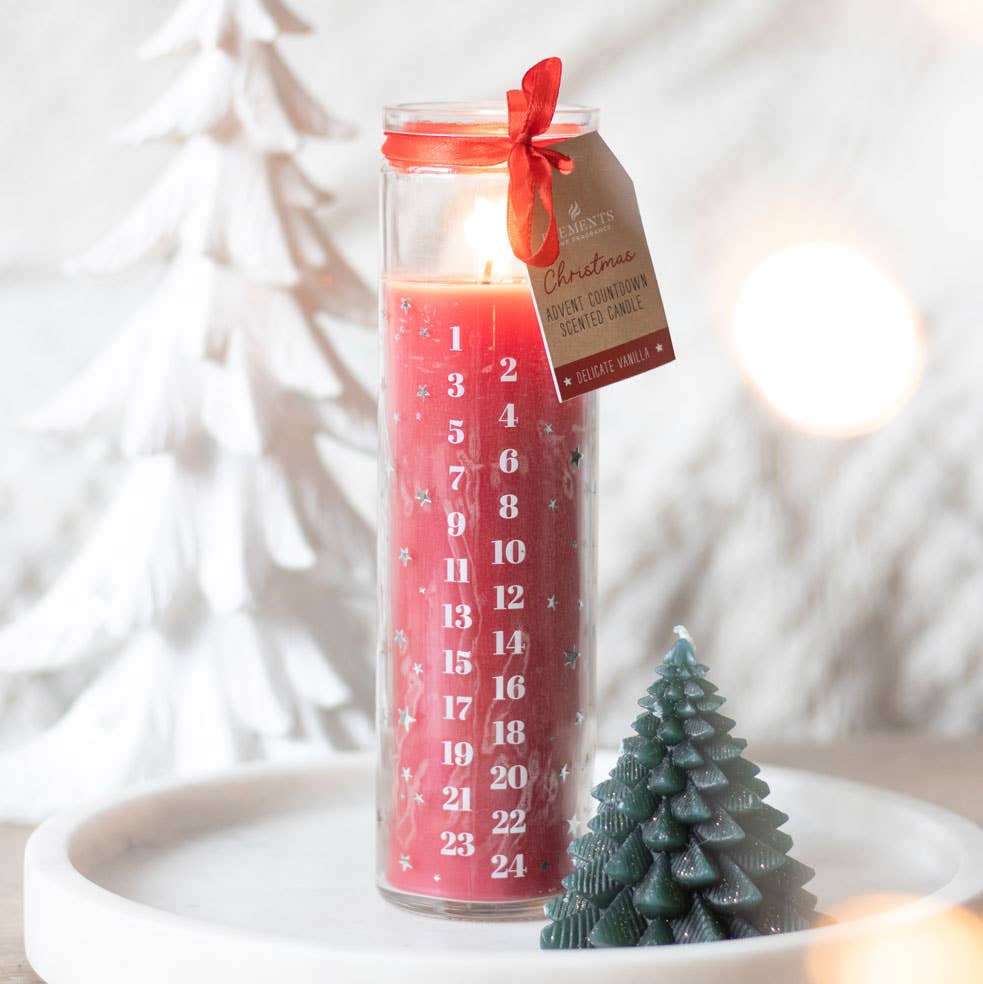 A red advent candle, known as the Red Vanilla Christmas Advent Tube Candle, crafted from vegan paraffin wax and adorned with numbers, is elegantly lit on a white plate. In the background, a green, tree-shaped candle with a delightful vanilla scent complements the scene alongside a white tree decoration.