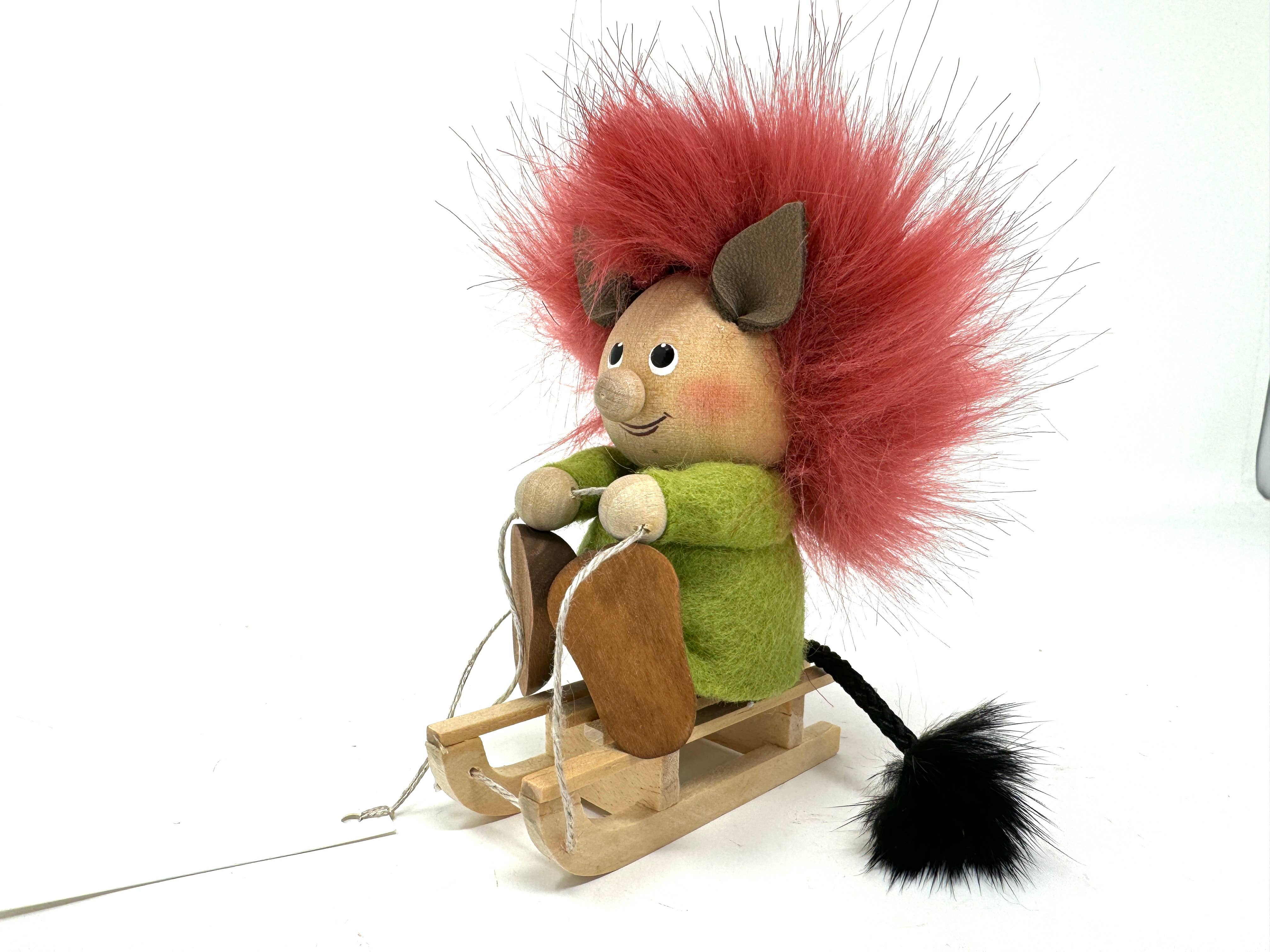 A handmade figurine named "Troll on a Sled" depicts a Scandinavian troll with red fluffy hair sitting on a wooden sled. This charming figure, ideal as a gift, features a smiling face, green outfit, brown boots, and a black furry tail.