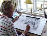An older person in a striped shirt meticulously sketches a detailed illustration of Sami Reindeer at their desk by the window, inspired by the Heidi Lange framed artwork titled Sami Reindeer (11x14) hanging nearby.