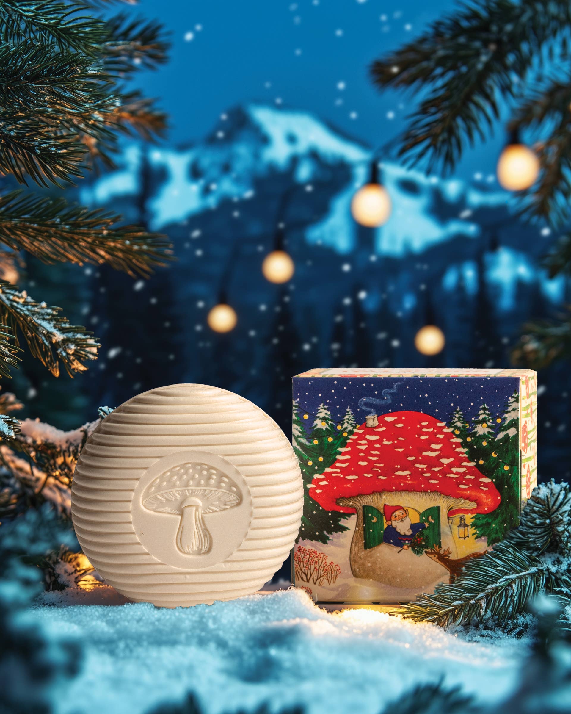 The Swedish Dream Gnome Soap, with its whimsical round mushroom design, is showcased in the snow next to an elegantly decorated box. Behind it, snow-covered branches and a mountain adorned with string lights create a backdrop that captures the enchanting essence of nature, reminiscent of Swedish pine forests where gnomes might reside.