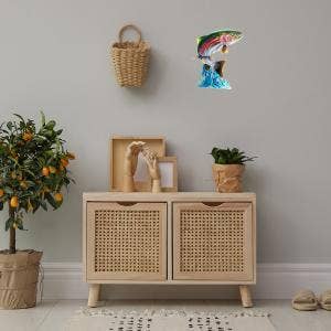 A wall-mounted piece named the "Jumping Trout Wall Art," crafted from American steel, is displayed above a cushioned storage bench, with plants and decorative items arranged beside it.