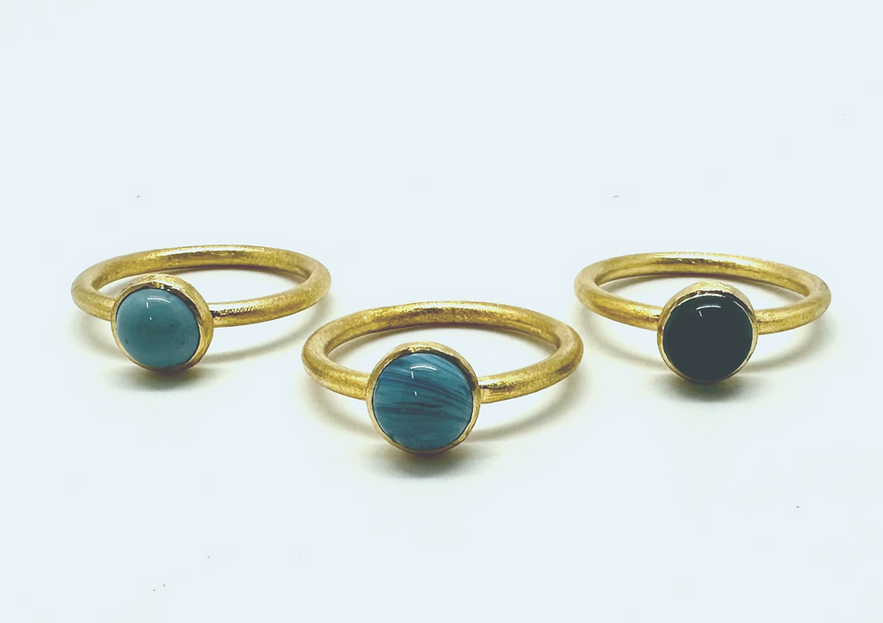 A trio of exquisite gold-plated rings, each with a round stone setting. The collection includes one with a vibrant turquoise stone, another named "Cirkel Gold" that showcases a unique Swedish Blue stone accented by distinctive lines, and the final piece adorned with an elegant solid black stone. All are beautifully displayed in sequence on an immaculate white background.