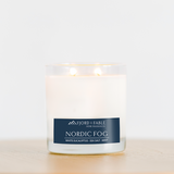 An 8oz Nordic Fog Candle by Fjord & Fable, featuring white eucalyptus, sea salt, and mint scent, sits in a glass jar on a wooden surface. This soy wax candle fills the air with a refreshing eucalyptus mint aroma.