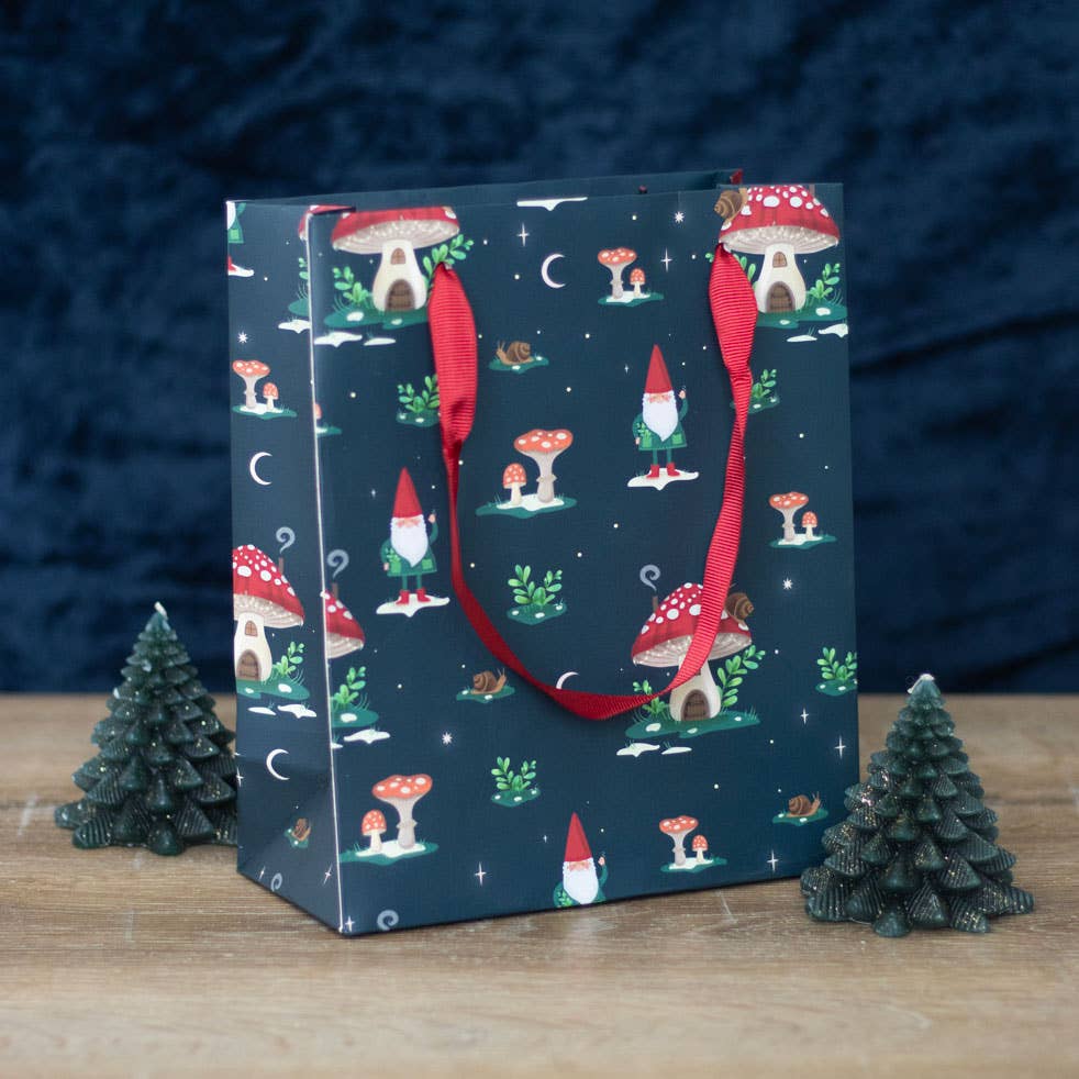 A 'Gnome Sweet Gnome' gift bag, inspired by a winter forest theme and featuring red handles, is adorned with a mushroom and gnome pattern. It is placed among small decorative tree figures on a wooden surface.