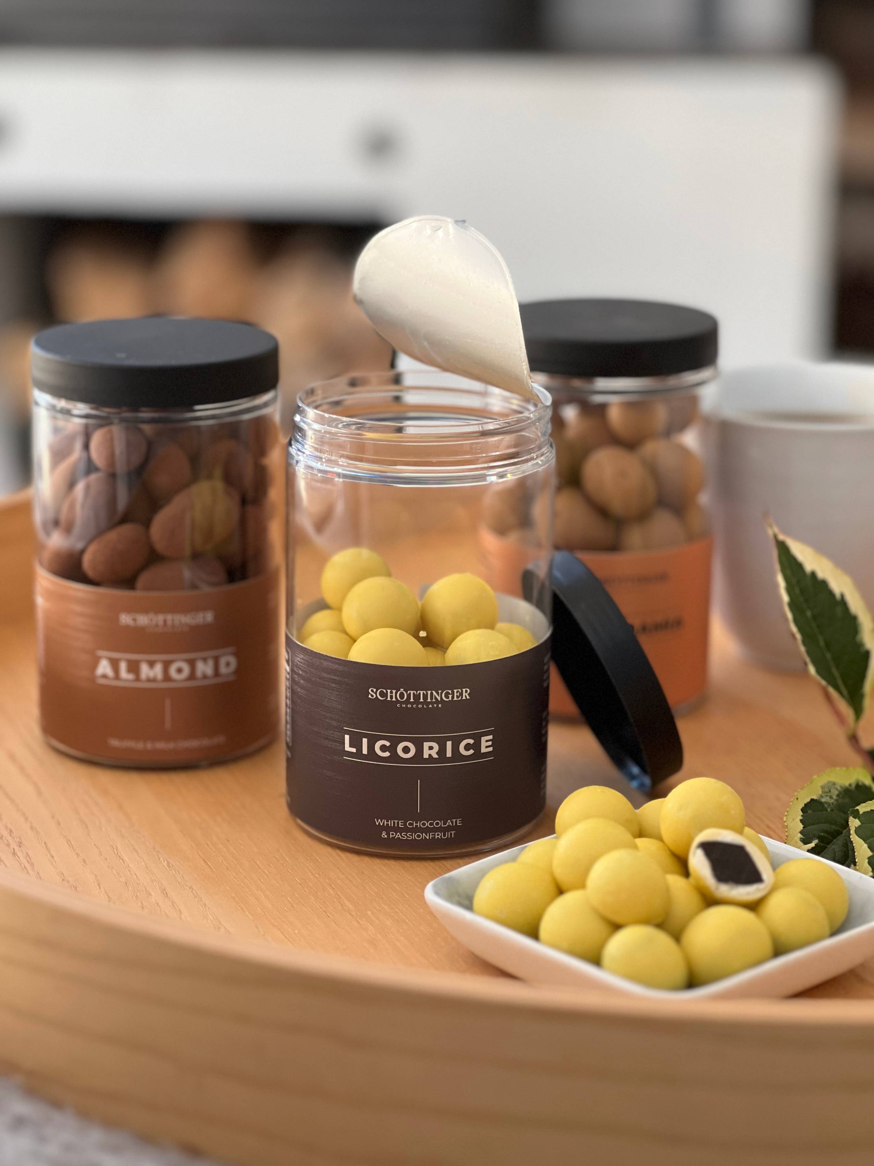 A wooden tray displays jars, including one for Schottinger Licorice with White Chocolate & Passionfruit. The opened jar reveals yellow candies, some on a plate with one cut open to show the creamy white chocolate filling inside.