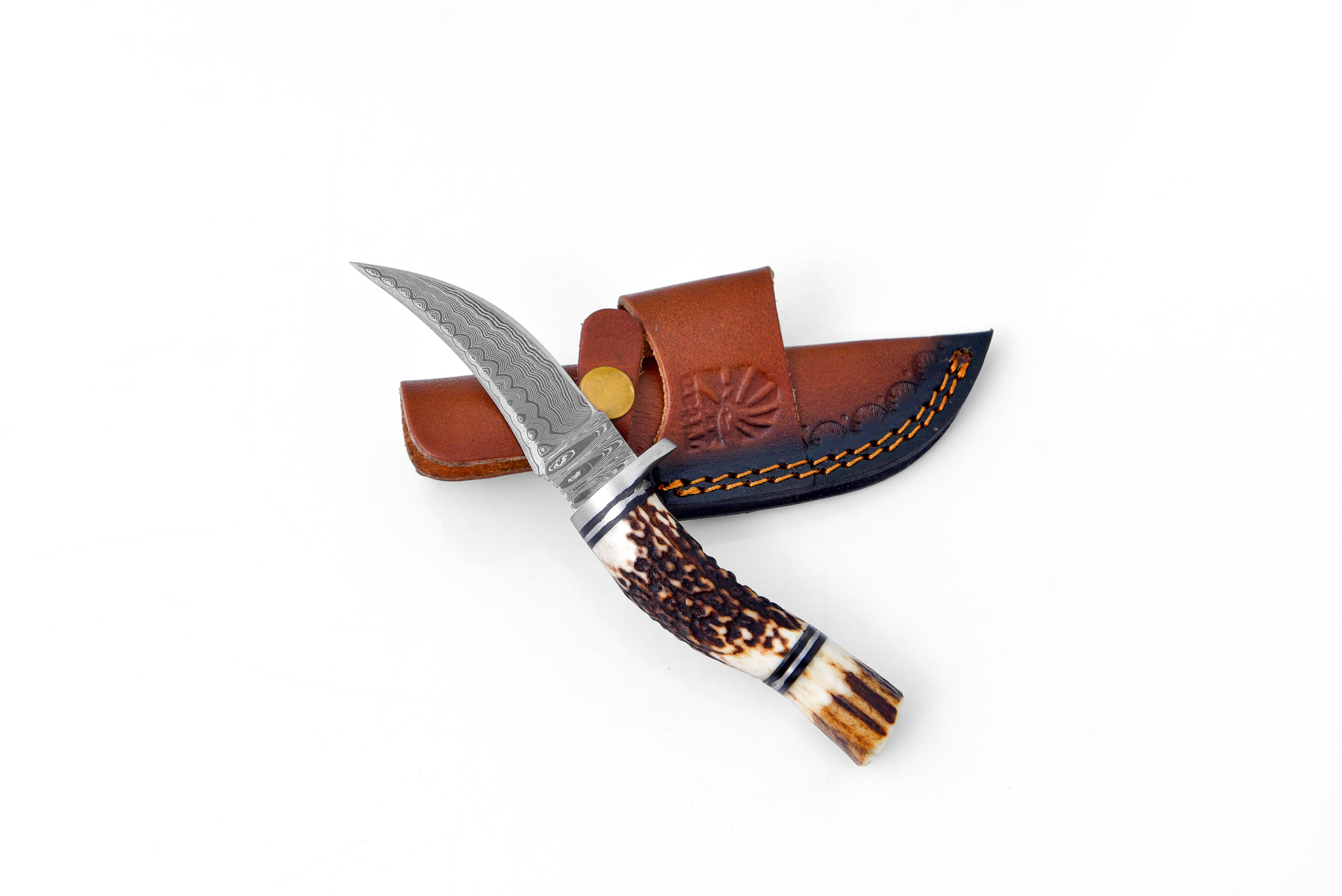 The Knife: Deer Horn Trailblazer Damascus by Titan features a hand-forged, patterned Damascus blade and a stag horn handle, resting on a brown leather sheath with decorative stitching.
