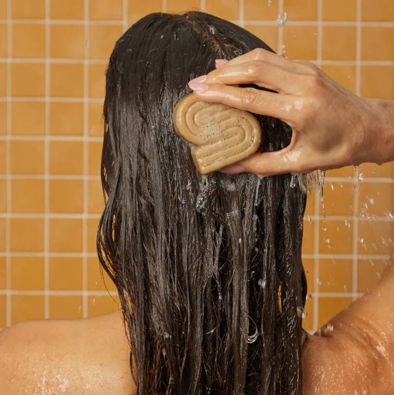 A person with wet, curly hair stands in a shower with orange tiles, washing their hair with the KITSCH Coconut Oil Shampoo Bar for Dry Damaged Hair, a brown S-shaped bar infused with moisturizing coconut oil.