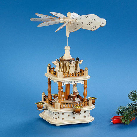 Experience the warmth of traditional holiday decor with the Marketplace24 Pyramid: Christmas Pyramid Nativity Square. Expertly crafted in Germany, this 13" tall piece features a manger scene on the bottom and four beautifully carved angels with horns on top, all set beneath a spinning fan. Shown against a blue background with a small fir branch and red candles nearby, this charming decoration captures the spirit of nativity celebrations and festive German craftsmanship.