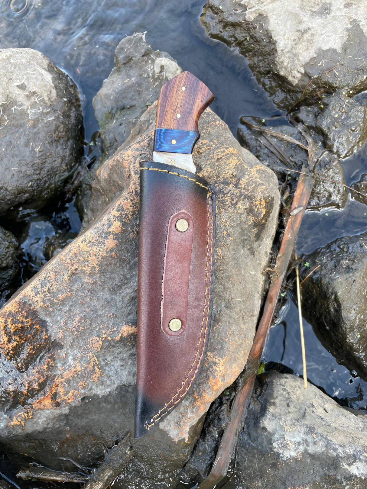 A Titan International Handmade High Carbon Damascus Steel Hunting Knife with a diamond wood and stag horn handle, in a leather sheath, rests on a large streamside rock among flowing water and scattered stones.