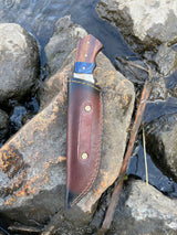 A Titan International Handmade High Carbon Damascus Steel Hunting Knife with a diamond wood and stag horn handle, in a leather sheath, rests on a large streamside rock among flowing water and scattered stones.