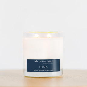 An 8oz Fjord & Fable Luna soy wax candle in a glass jar with 2 wicks and 45+ hour burn time rests on wood. The label features notes of violet, saffron, vetiver, and oud for a unique home fragrance experience.