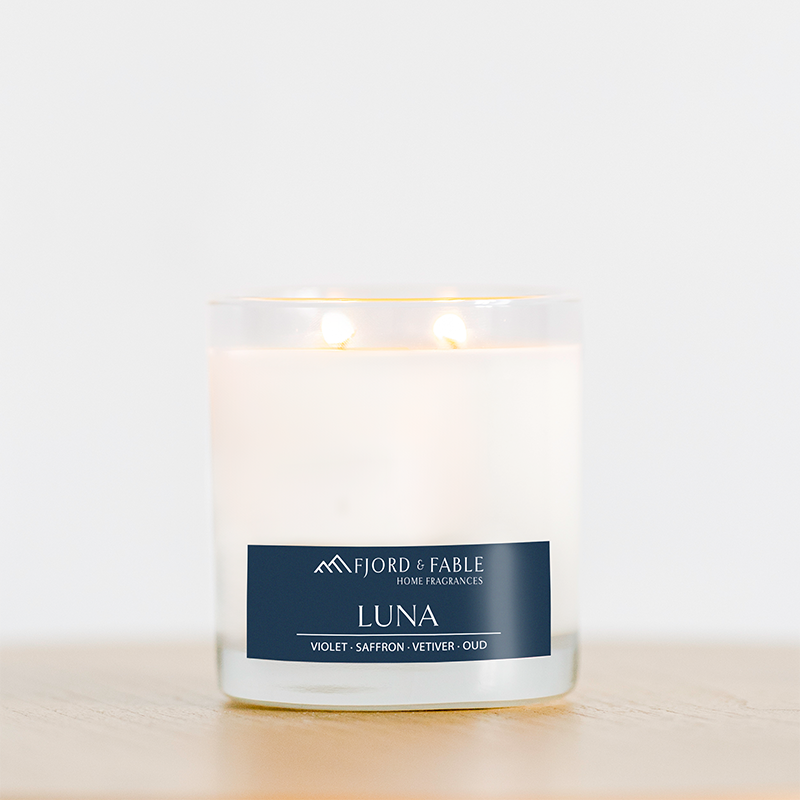 A white vegan Luna Candle: 4oz with three lit wicks in a glass jar labeled Fjord & Fable, featuring notes of Violet, Saffron, Vetiver, and Oud, sits on a light wooden surface against a plain background.
