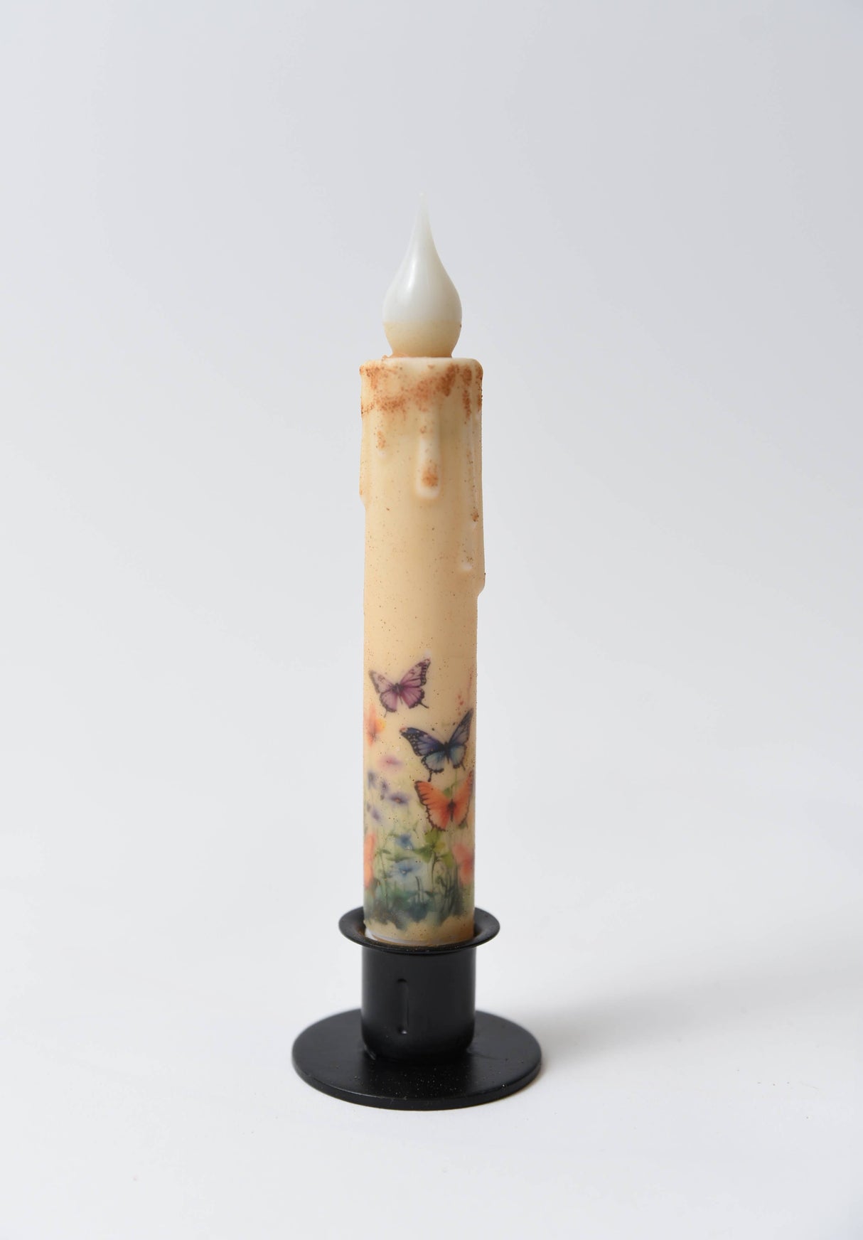 The Candle: Butterfly Panorama LED Timer Taper Candle: Top Dusted is a battery-operated LED taper with a faux wax finish, featuring butterfly and floral motifs in a black holder against a plain white background.