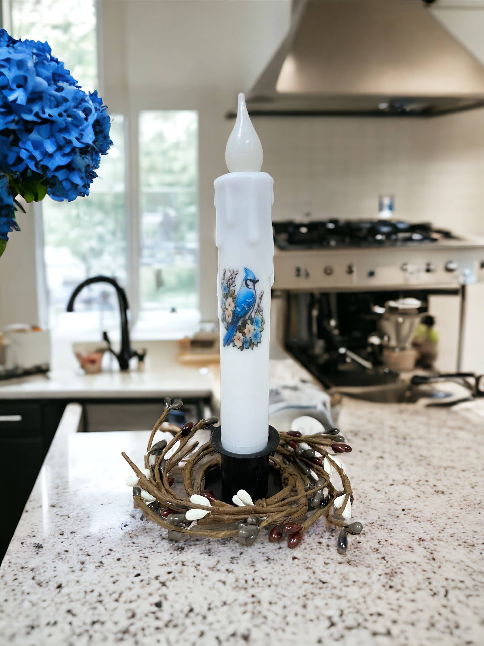 The kitchen counter features a Blue Jay Spring Bird battery-operated white candle with a timer, enveloped by twigs and small colored stones. To the left are vibrant blue hydrangeas, while kitchen appliances subtly backdrop the scene.